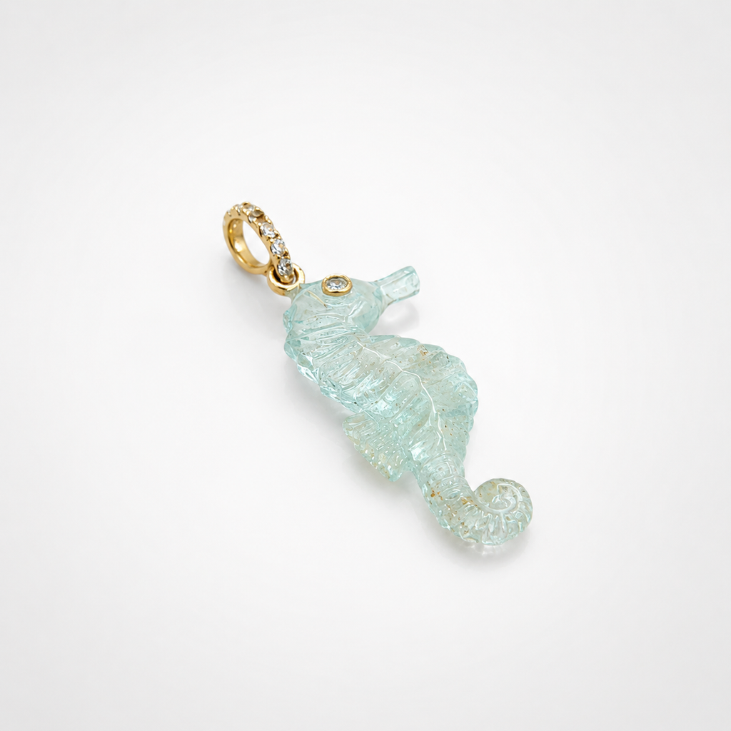 Hand-carved aquamarine seahorse pendant shown at diagonal angle, with 14k yellow gold open-ring pavé bail set with white topaz accents and bezel-set white topaz eye, translucent pale blue gemstone with natural inclusions and detailed scale carving