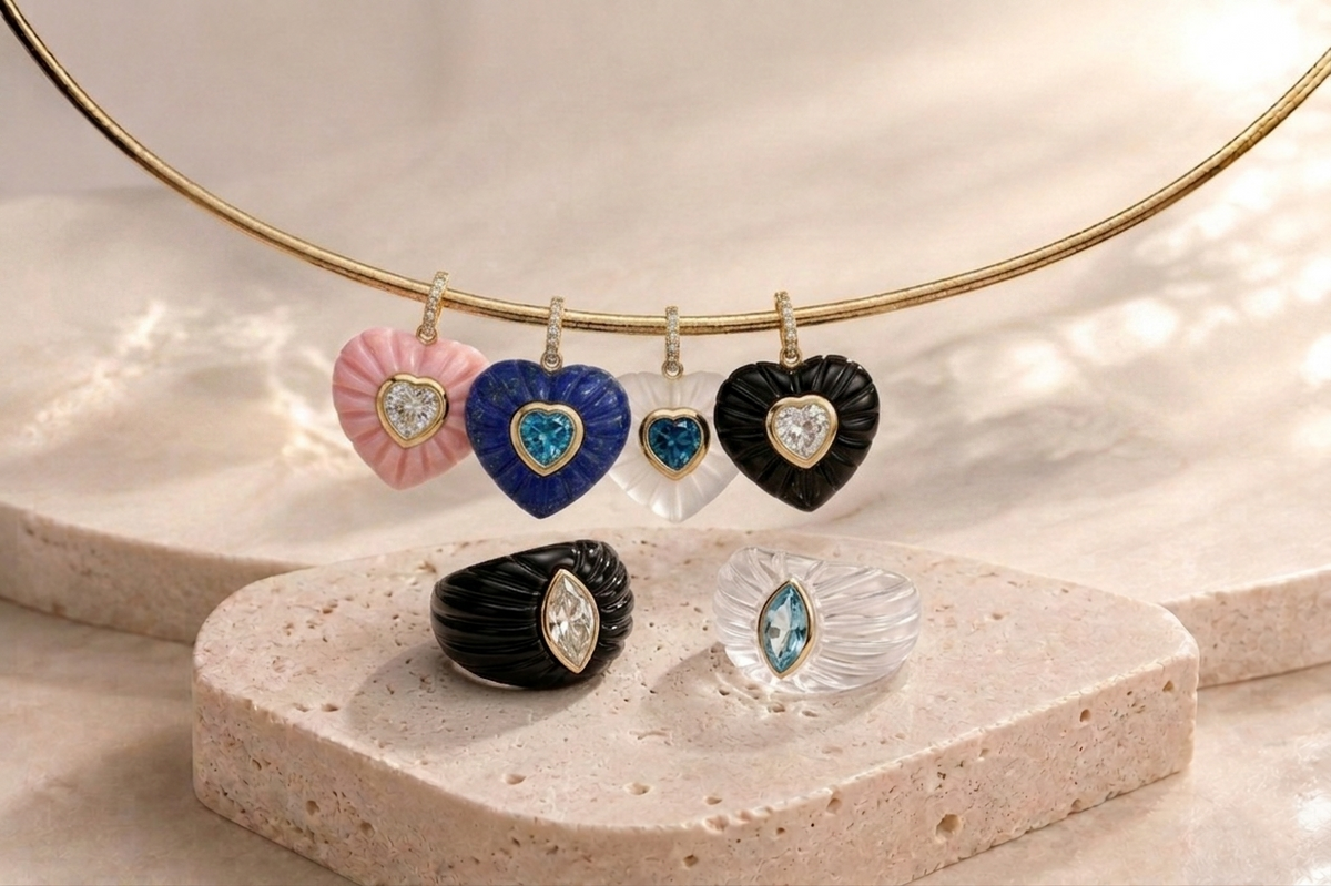 Editorial flat lay of Aurelia heart pendant collection hanging on 14k yellow gold pavé bangle — pink opal with white topaz, lapis lazuli with Swiss blue topaz, frosted rock crystal with Swiss blue topaz, and black onyx with white topaz — alongside black onyx fluted ring with marquise white topaz and frosted rock crystal fluted ring with marquise Swiss blue topaz, styled on travertine stone surface