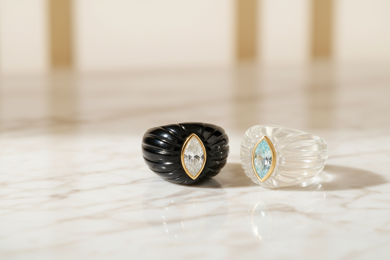 Black onyx fluted dome ring with marquise white topaz gold bezel alongside a clear rock crystal fluted dome ring with marquise blue topaz gold bezel on marble surface